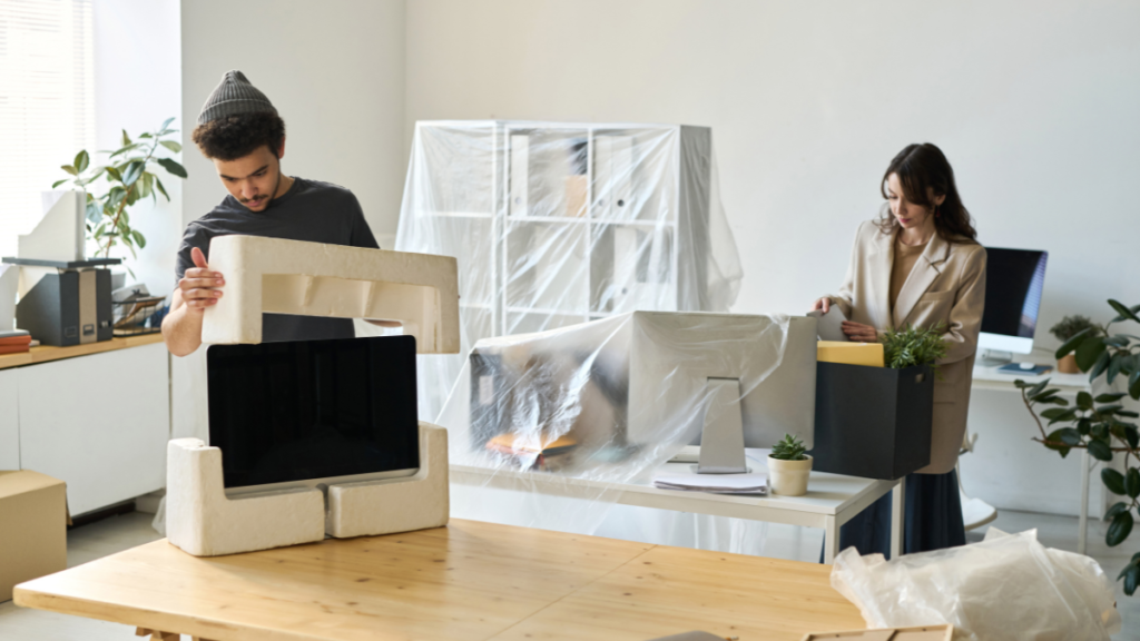 Male packing computer, female packing in a box. Wrapped office furniture in plastic